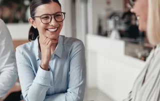 Two women, both wearing glasses, are sitting and smiling at each other across a table. One has a notepad and pen in front of her. They appear to be in a modern office environment.