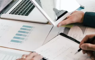 Two people working at a desk with a laptop, notepad, and documents featuring charts and graphs.
