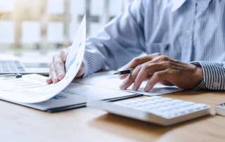 A person in a striped shirt reviews documents at a desk with a calculator, pen, laptop, and papers under daylight.