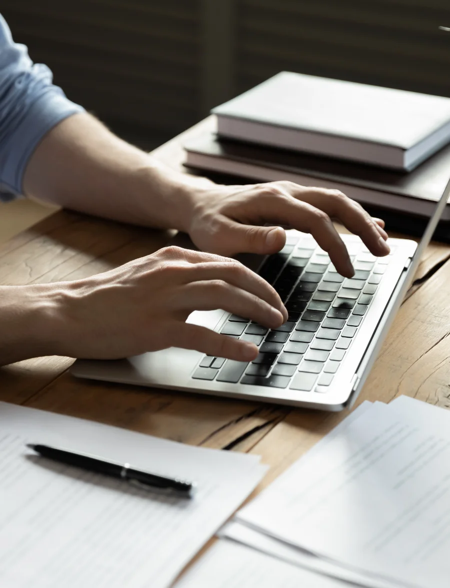 A person types on a laptop at a wooden desk with documents, a pen, and stacked books nearby.