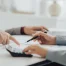 Two people in business attire discuss real estate tax graphs and data at a desk; one person points at a document while the other uses a calculator and holds a pen.