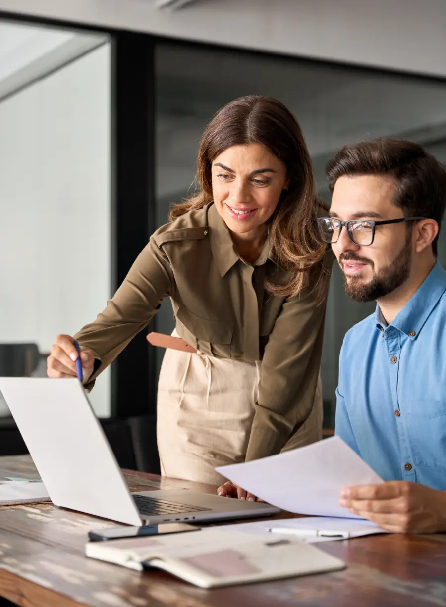 Two colleagues working together at a desk, one pointing at a laptop screen displaying real estate tax data while the other holds a document.